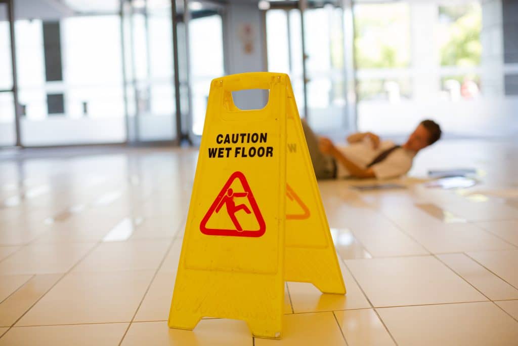Slip and fall injuries scene in a commercial building, with a yellow caution wet floor sign in focus and a person lying on the ground in the background.
